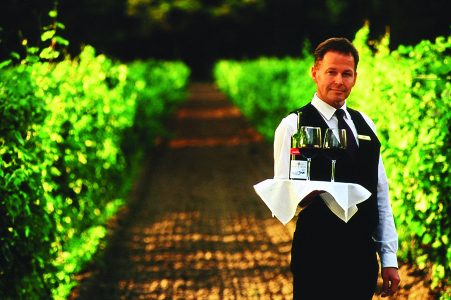A well-dressed man standing in a vineyard holding a tray with a bottle of red wine and two glasses