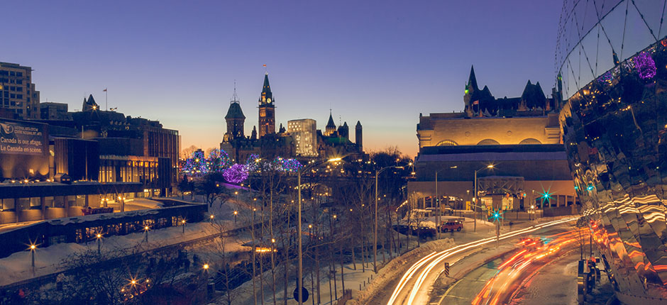 Night shot of Ottawa along the Rideau Canal, showcasing the traditional architecture of the capital building next to a modern, curved mirrored building with interlocking triangular windows